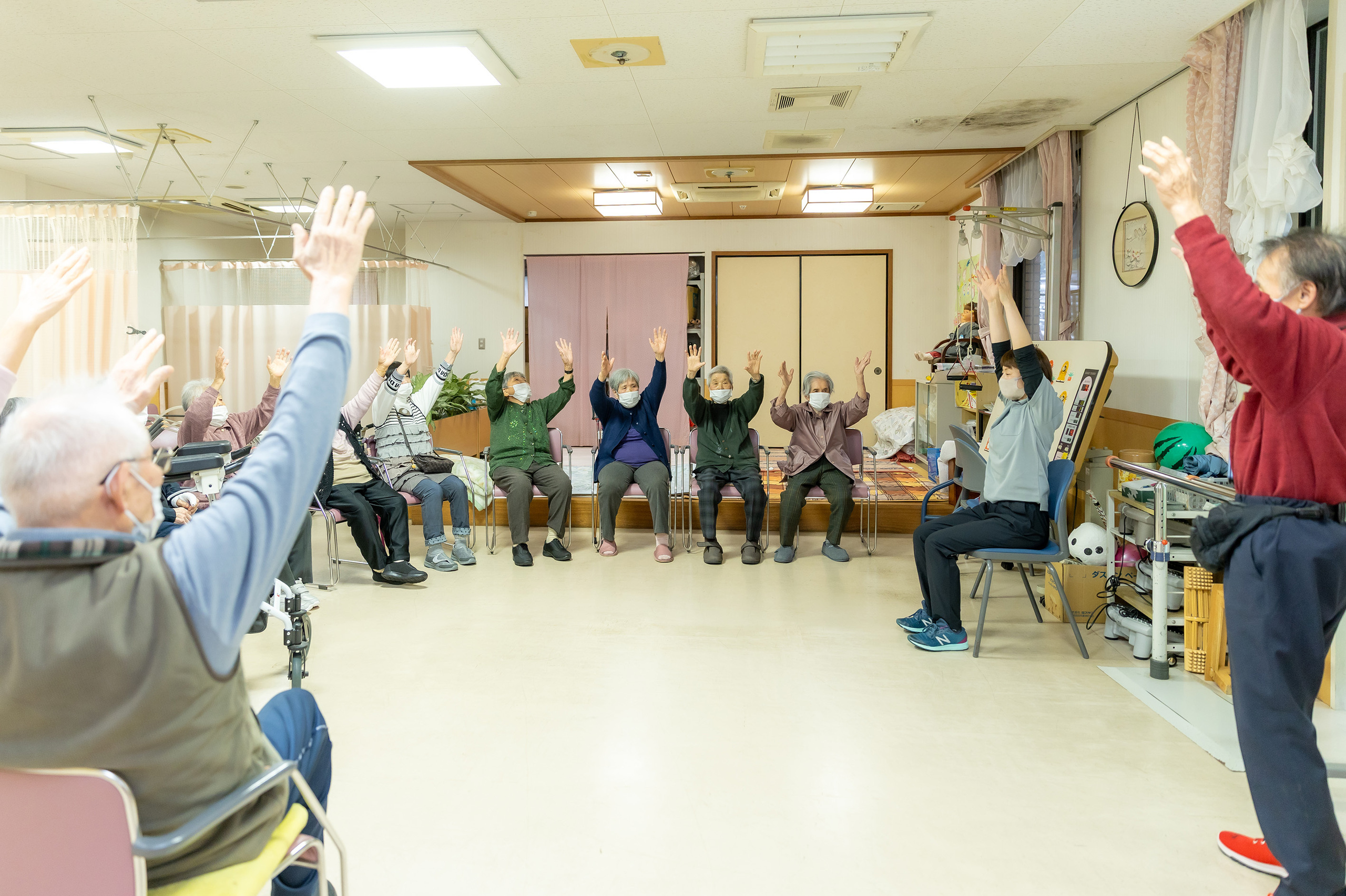 介護施設での集団体操・レクリエーション。入居者と職員が腕を上げて運動している。